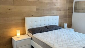Een slaapkamer met een wit bed, een nachtkastje met een lamp en een houten wand.