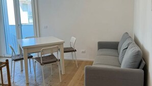 Een compacte, moderne woonkamer met een grijze bank, een witte eettafel met twee stoelen en een openslaande deur naar een balkon.