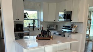A modern kitchen with white cabinetry, a central island with stools, a stainless steel refrigerator, a microwave, a toaster oven, and a coffee maker. - Treetop getaway on a secluded Island with gulf coast beaches  (Captiva)