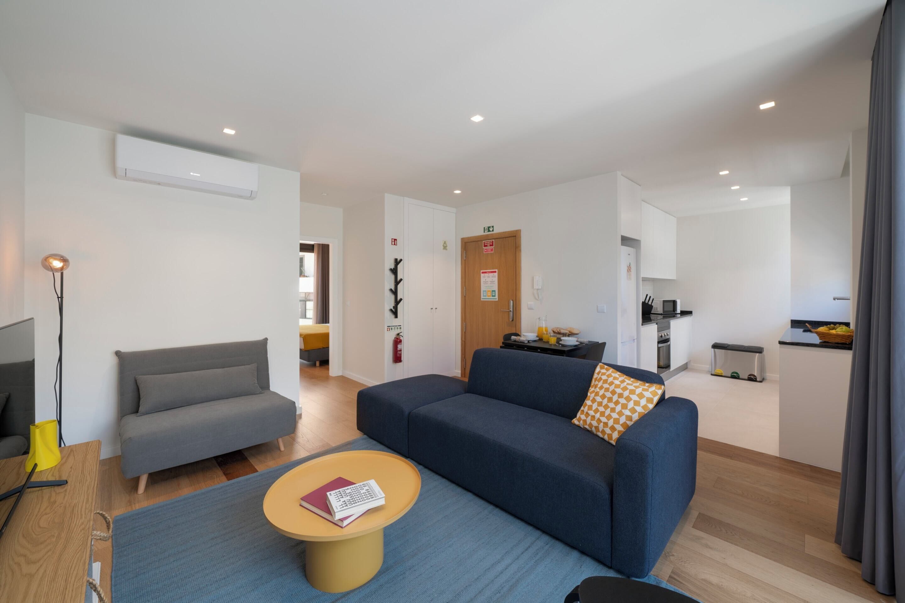 A modern living room with a sofa, a small coffee table, and a TV. There is a kitchen area in the background.
