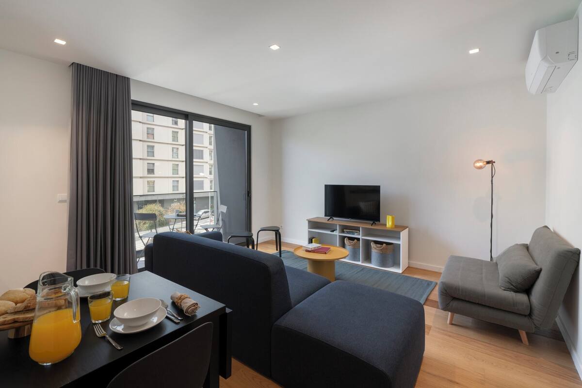 A modern living room with a sofa, a dining table with breakfast, a TV, and a large window.