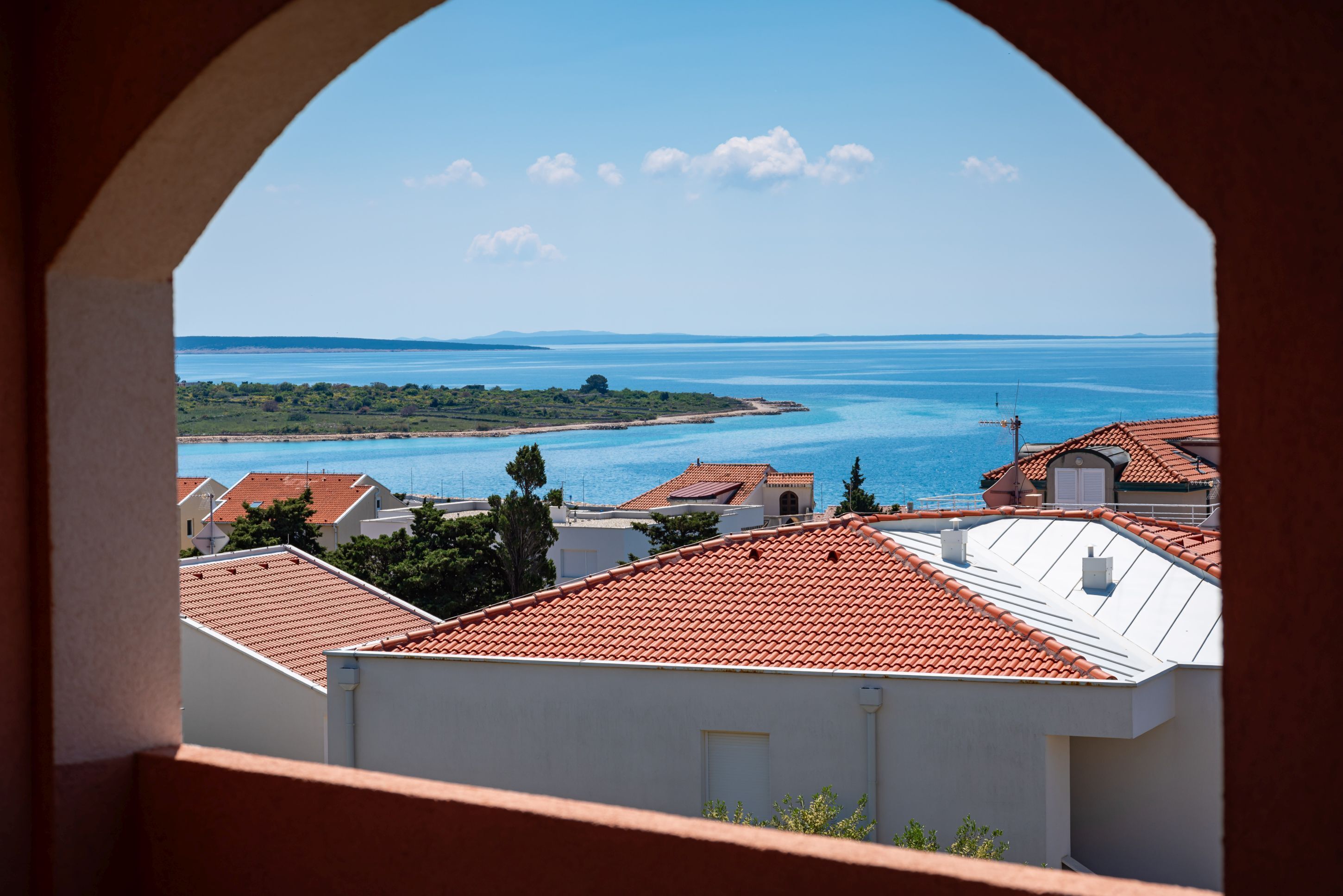 A coastal view through an archway, featuring buildings with red-tiled roofs and a clear blue sea.