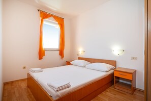 A bedroom with a wooden bed, a nightstand, a desk, a window with orange curtains, and wall-mounted lights. - Comfortable Apartments Malina A4 br 3 (Novalja)