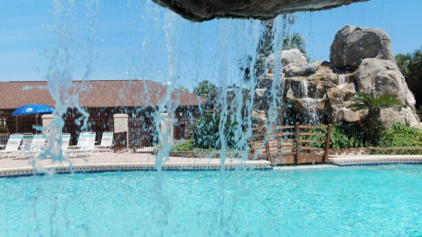 Une piscine d’un bleu éclatant, agrémentée d’une cascade, de chaises longues et, au fond, d’un bâtiment dont le toit est recouvert de tuiles.