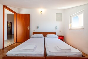 A room with two beds, a wooden wardrobe, a nightstand, a wall-mounted light, and a window.