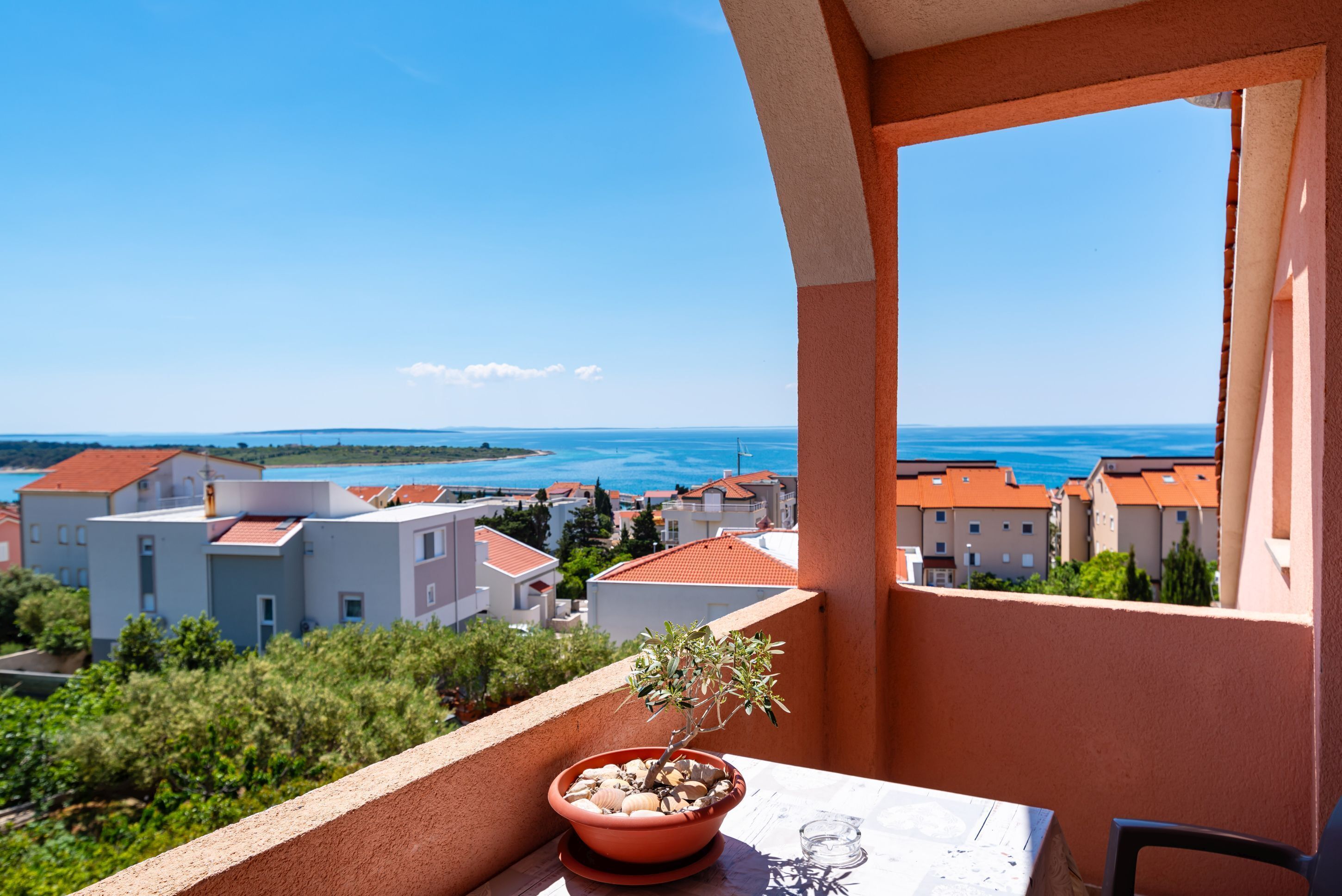 A balcony with a potted plant, a table, and a clear view of a coastal town.