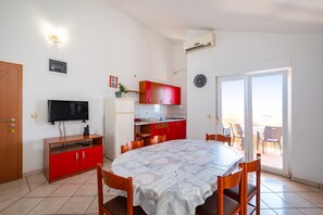 Un ambiente compatto con tavolo e sedie da pranzo, televisione, angolo cottura e una porta-finestra scorrevole verso un’area esterna.