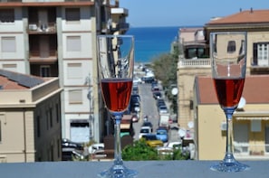 Two wine glasses on a balcony with a view of a coastal street and buildings.