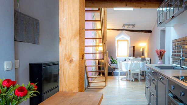Une cuisine moderne avec des poutres en bois, un coin repas avec une table et des chaises blanches, et une vue sur une chambre avec une fenêtre.