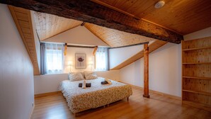 Une chambre avec un plafond en pente, des poutres en bois, un lit recouvert d’une couvre-lit à motifs, deux lampes de chevet, une fenêtre avec des rideaux et une bibliothèque en bois.