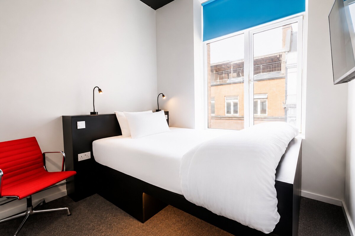 A hotel room with a bed, a red chair, a desk with a lamp, and a window with a view of buildings.