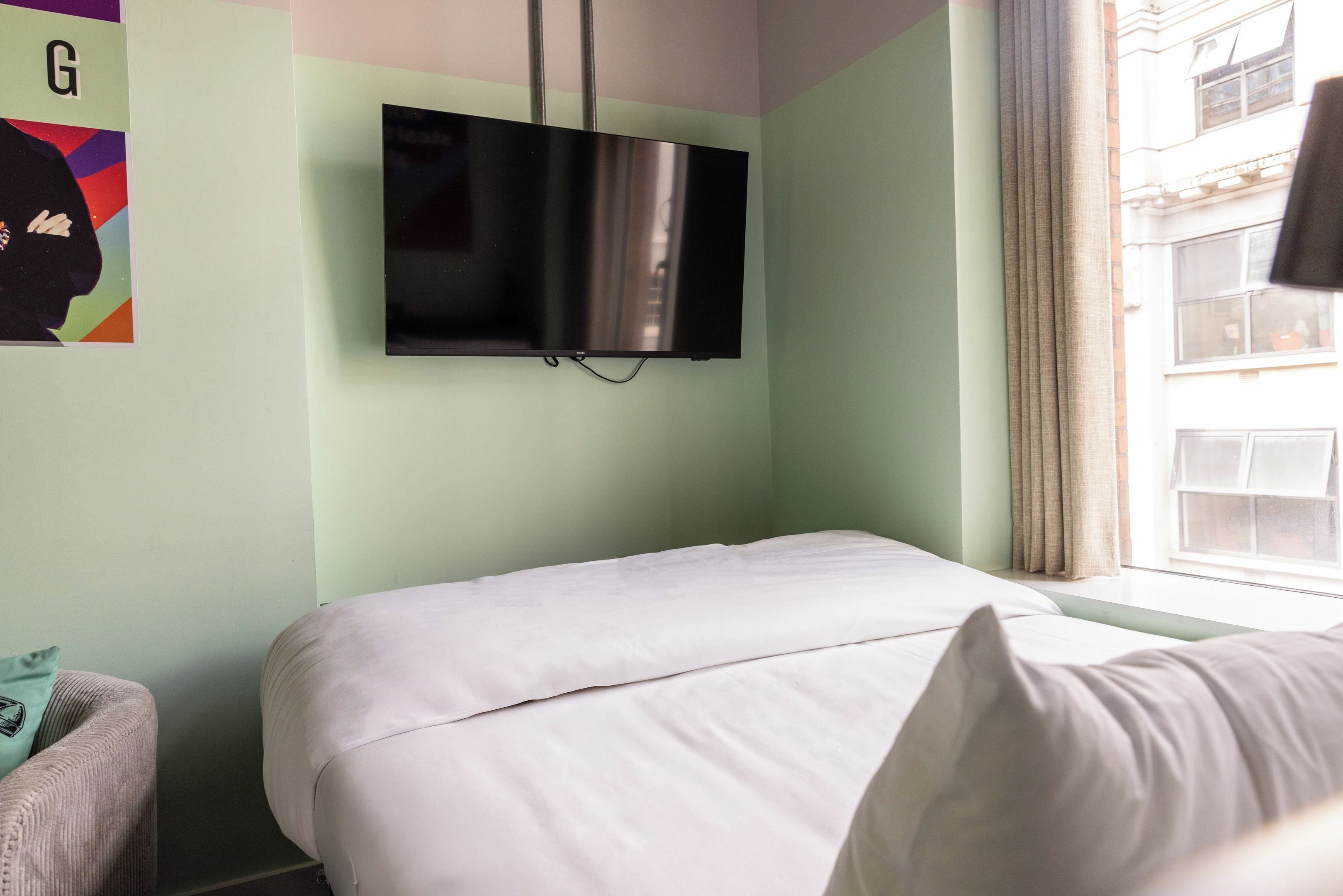 Ein Schlafzimmer mit einem Bett, einem an der Wand befestigten Fernseher und einem Fenster mit Vorhängen.