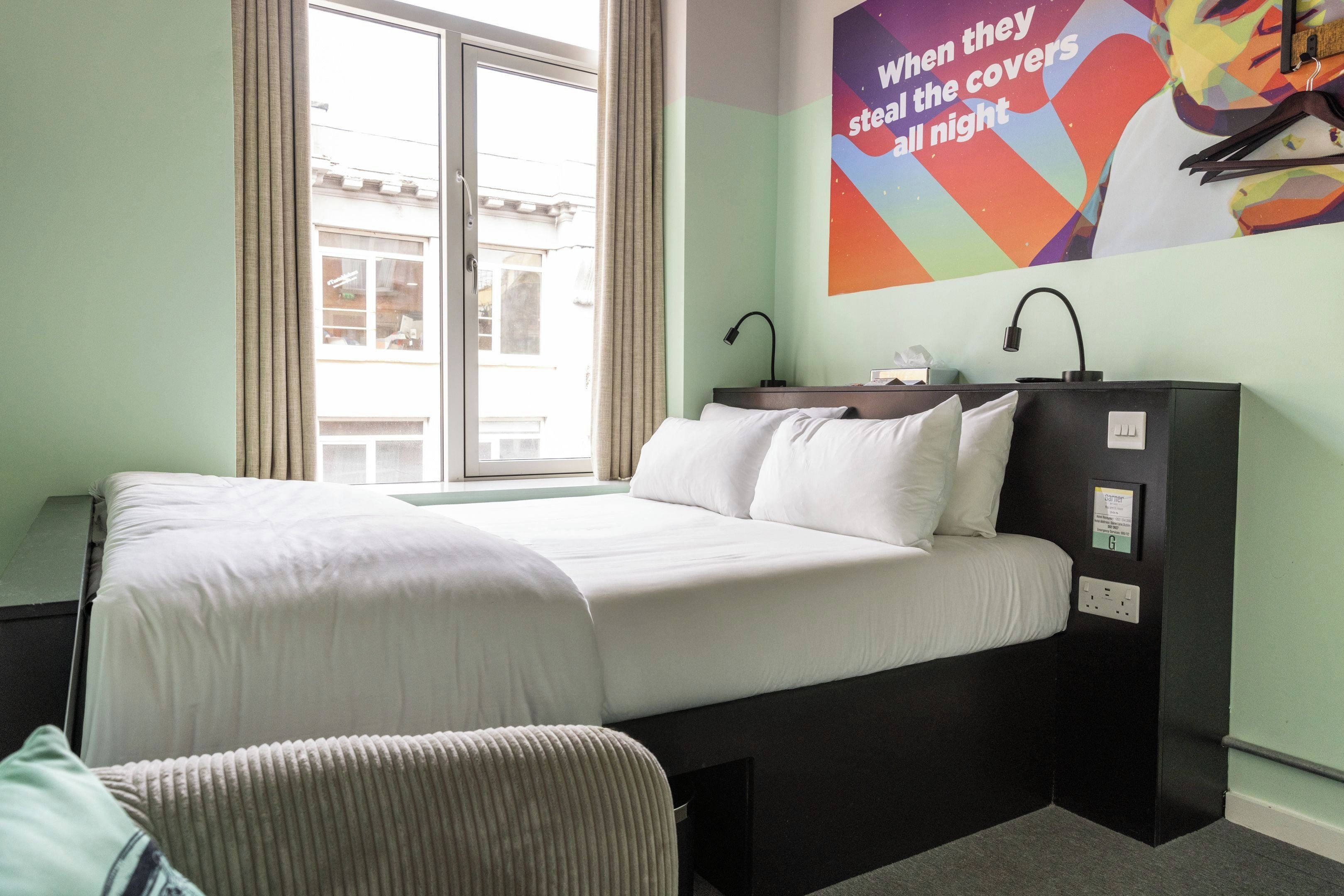 A hotel room with a bed, a desk, a chair, a window with curtains, and a wall with a colorful poster.