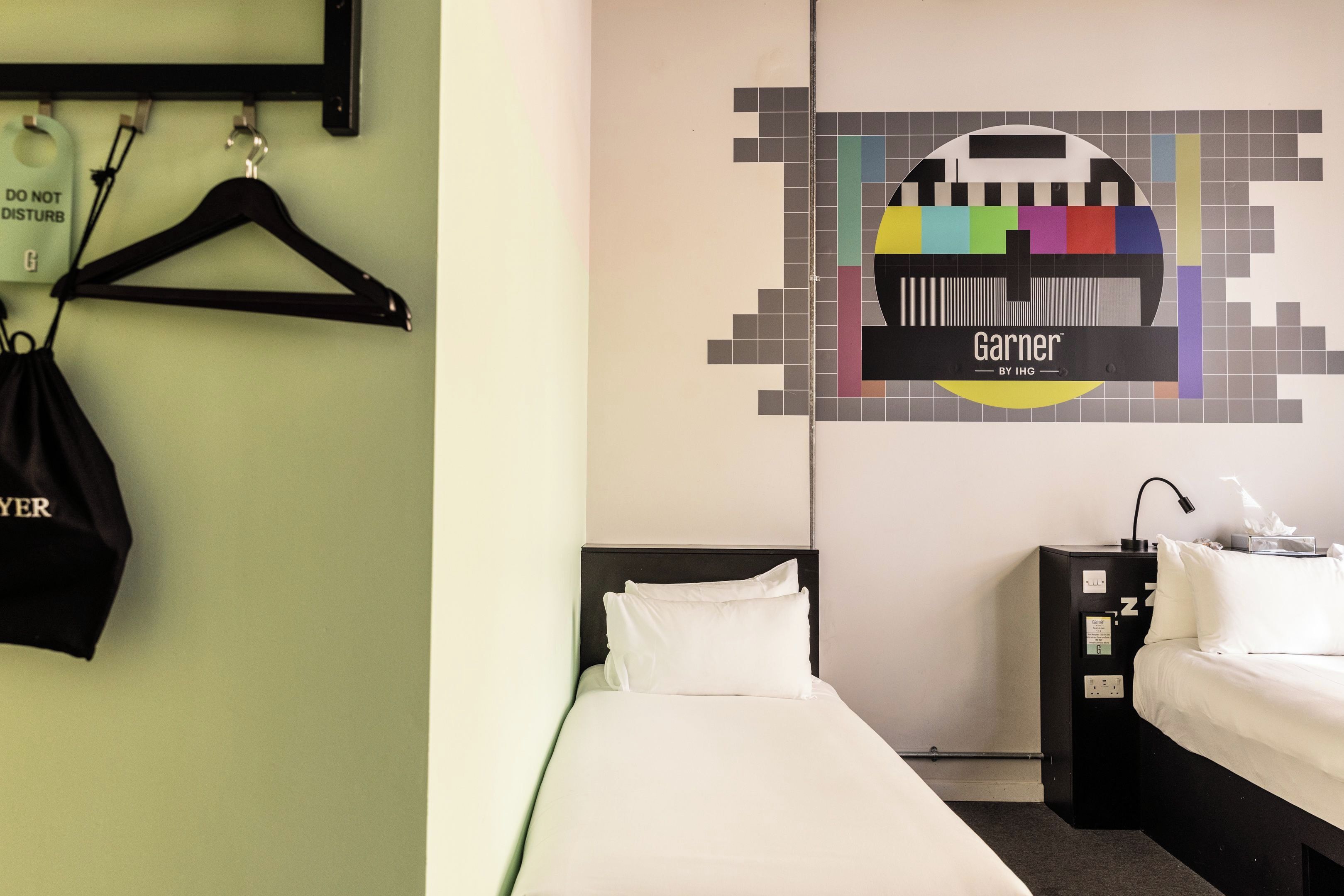 A hotel room with a bed, a nightstand, a lamp, and a wall with a pixelated Garner logo.