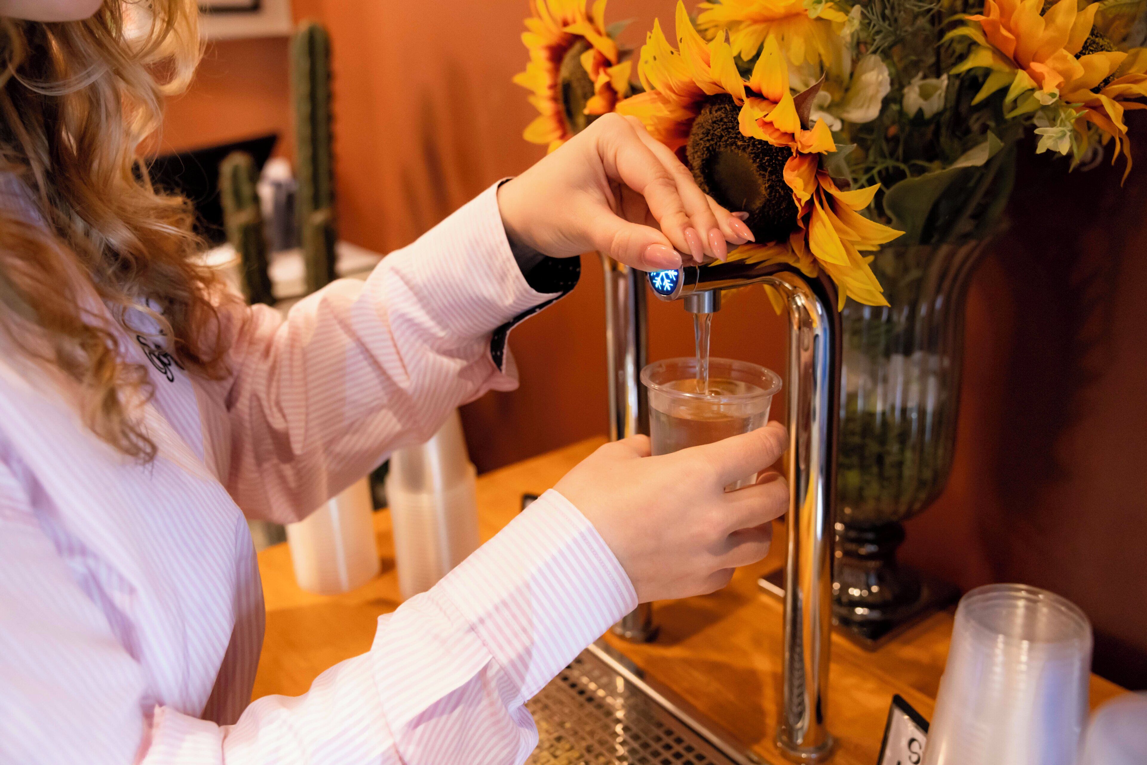 Una persona llenando un vaso con agua de un grifo, un jarrón con girasoles, y una zona de bar con mostrador y taburetes.