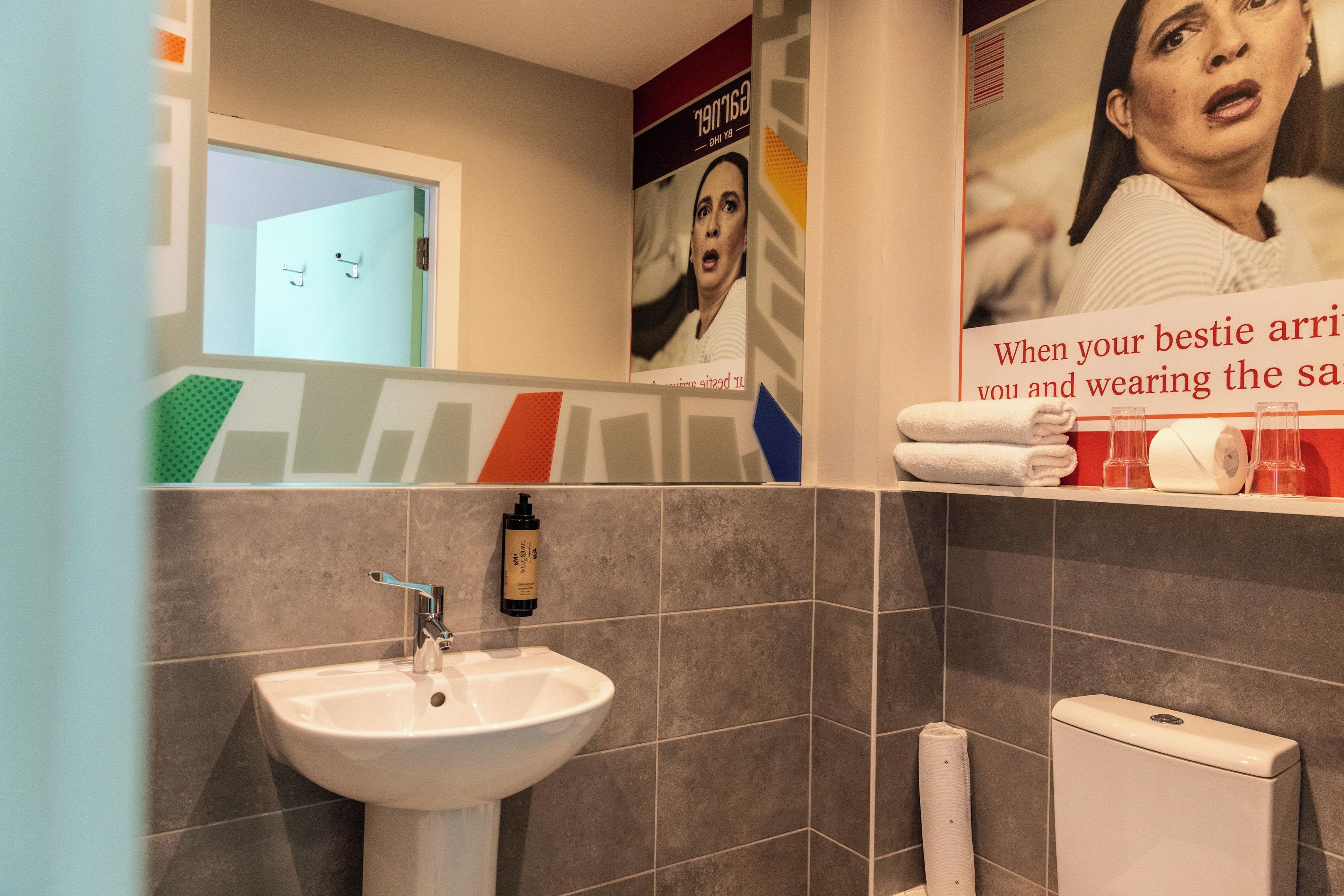 A bathroom with a white sink, a toilet, and a shower area. There are posters on the wall and a towel rack with towels.
