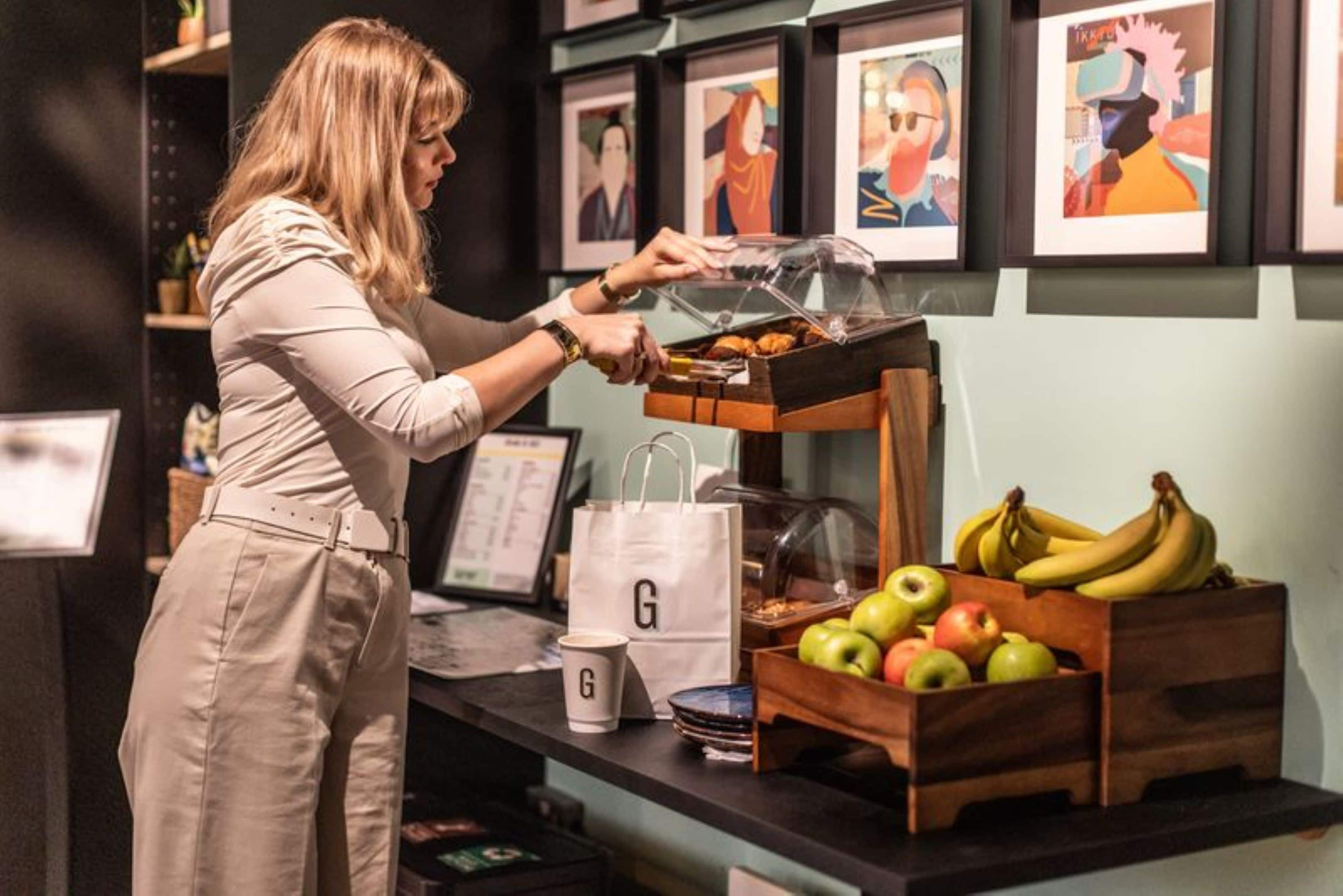 A person serving food from a glass display case, with a fruit basket and a white bag labeled 'G' on the counter.