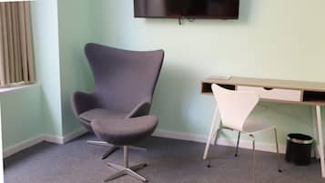 A room with a grey upholstered chair, a white chair, a wooden desk with drawers, a television mounted on the wall, and a black trash can.
