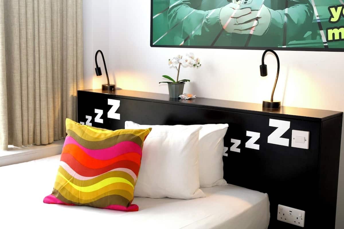 A modern hotel room with a black headboard, a bed with white bedding, a colorful pillow, and a small potted orchid.
