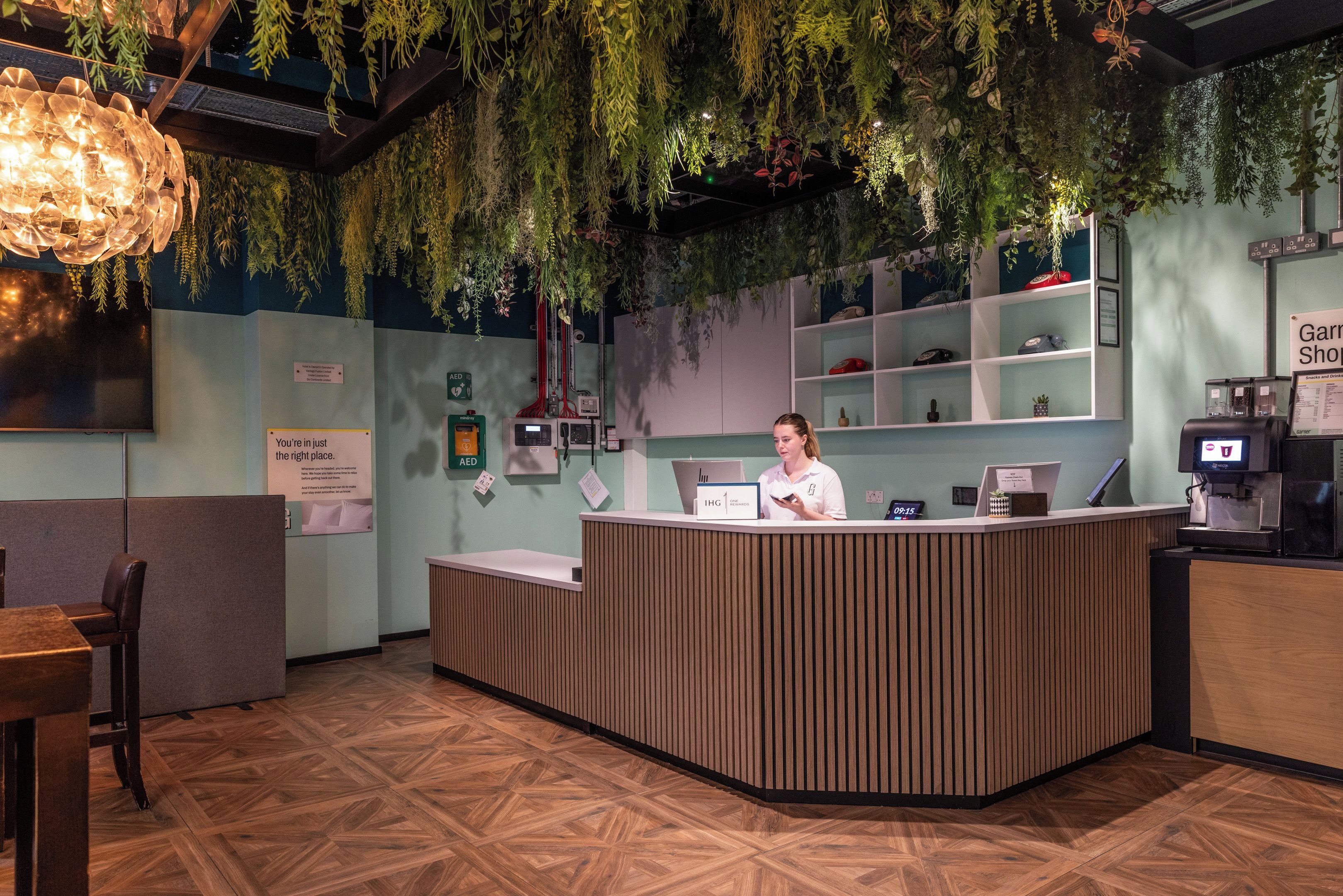 A reception desk with a woman standing behind it, surrounded by indoor plants, a digital clock, and a menu board.