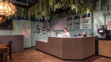 A reception desk with a woman standing behind it, surrounded by indoor plants, a digital clock, and a menu board.
