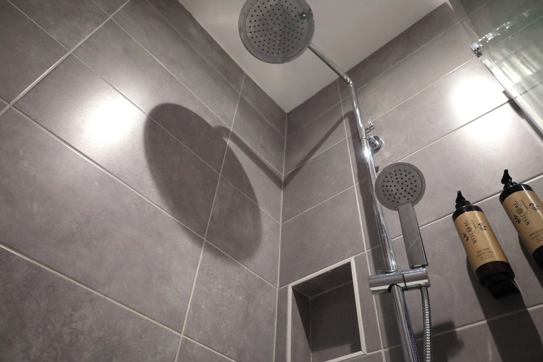 Un espace douche avec des carreaux gris, une douchette à main et deux flacons de lotion pour le corps fixés au mur.
