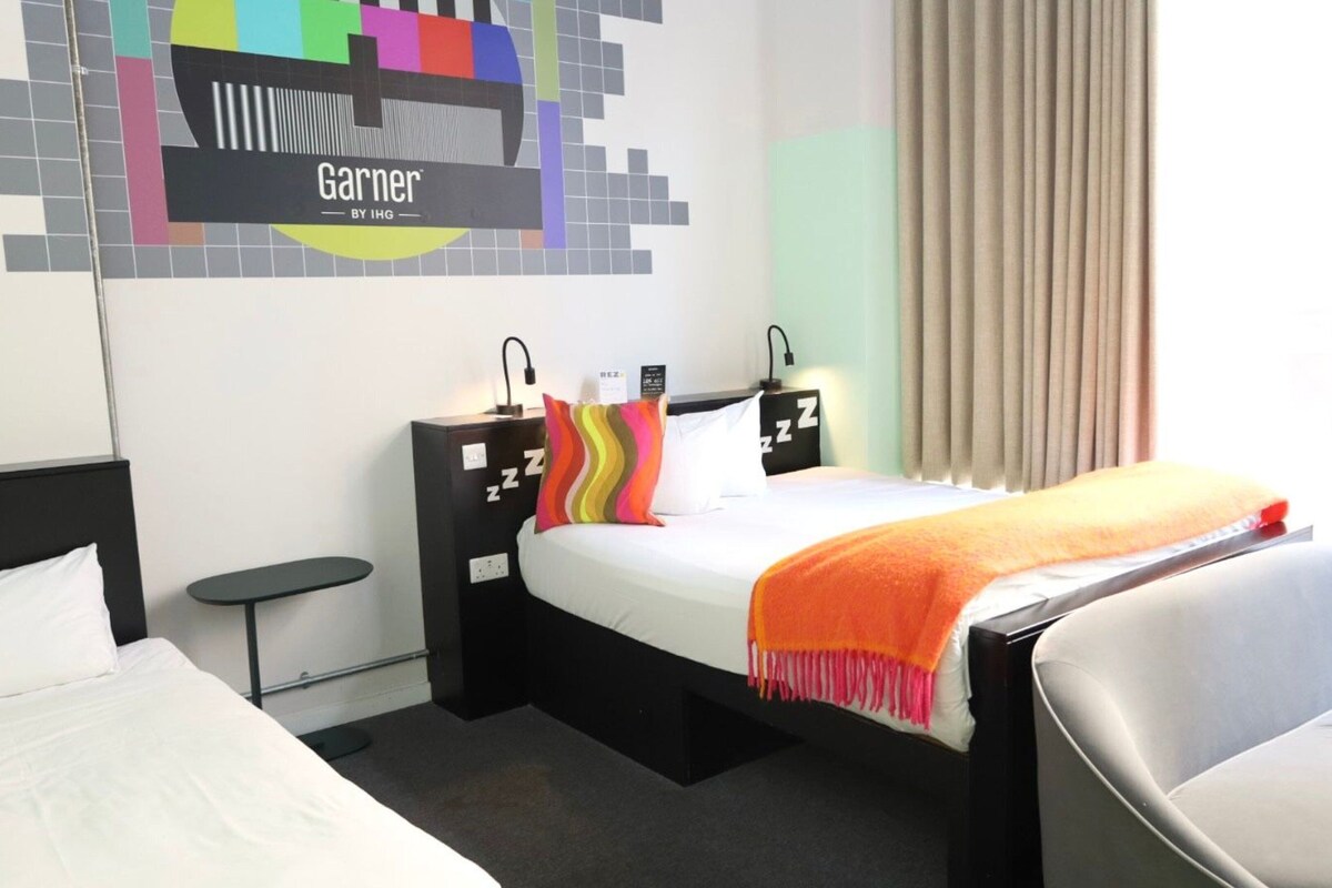 Une chambre d’hôtel comprenant un lit, des tables de chevet, une chaise, un téléviseur fixé au mur et une fenêtre avec des rideaux.