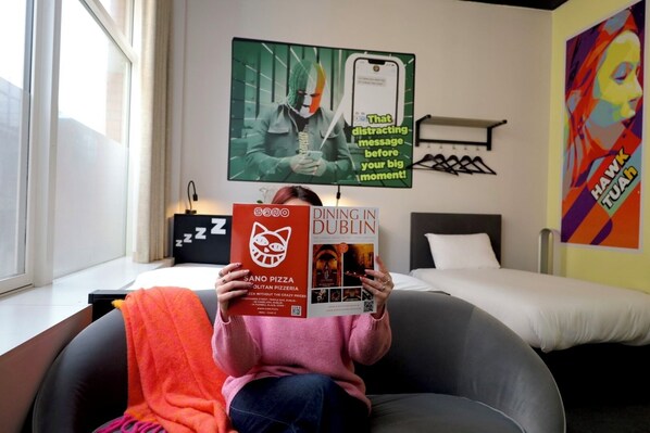 Una persona seduta su una sedia, che legge un libro intitolato "DINING IN DUBLIN" in una camera d'albergo con un letto, una finestra e un quadro appeso al muro.