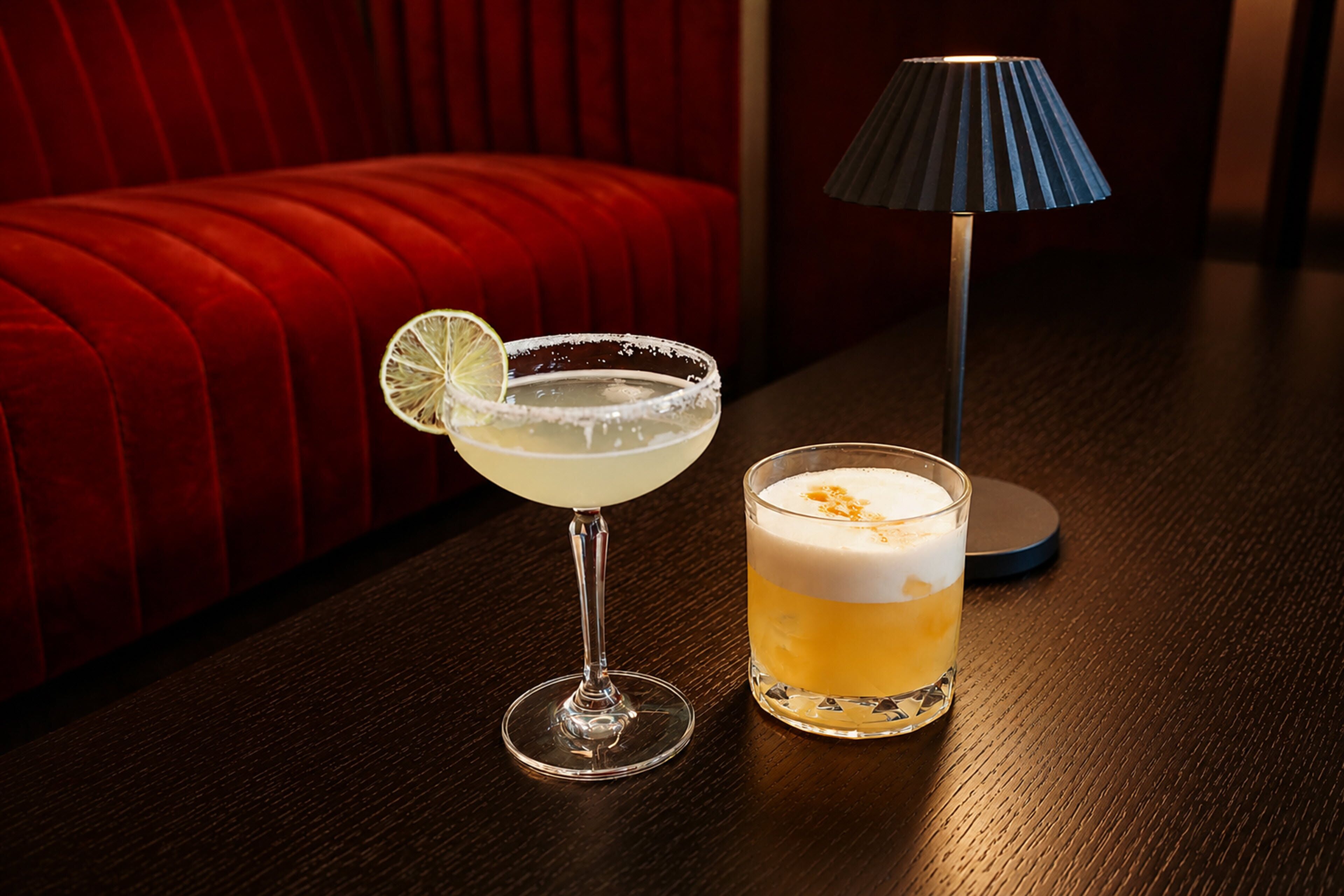 Un verre à cocktail avec un bord décoré, une tranche de citron en guise de garniture, et une boisson à plusieurs couches à côté d’un grand verre dont le bord est également décoré avec du sucre.