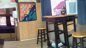 Ein Zimmer mit einem Holztisch, zwei Hocker, einem Poster an der Wand und einem Bücherwagen.