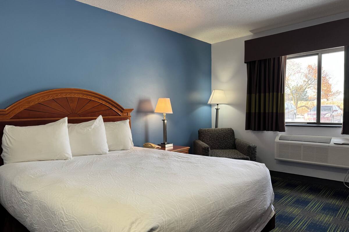 A hotel room with a large bed, two bedside tables, a chair, a window with curtains, and an air conditioning unit.