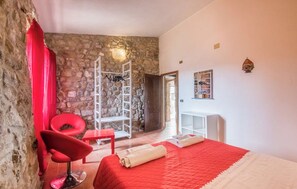 Une chambre avec un mur en pierre, une chaise rouge, un lit recouvert d’une couverture rouge, une table de chevet blanche et une étagère blanche.