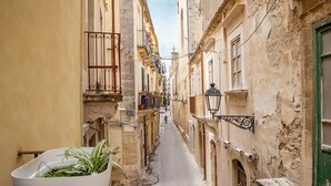 A narrow street with buildings on either side, a white trash bin with plants, and a person walking in the distance.