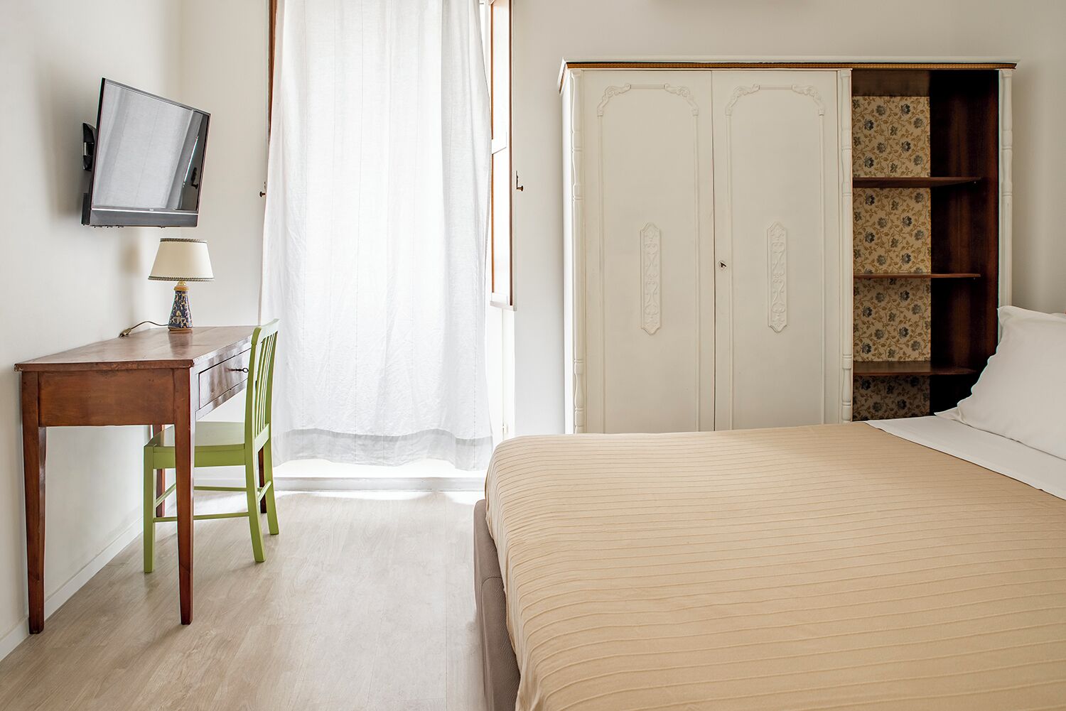 A bedroom with a bed, a desk with a lamp, a chair, a wardrobe, and a television on the wall.