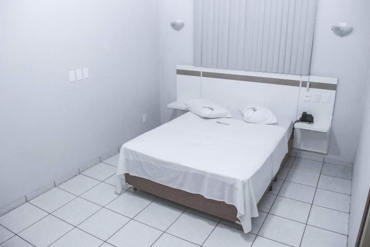 A clean, white hotel room with a single bed, a bedside table, and a small window with blinds.