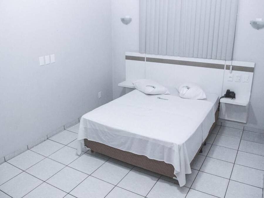 Quarto de hotel branco e limpo com uma cama de solteiro, uma mesinha de cabeceira e uma pequena janela com persianas.