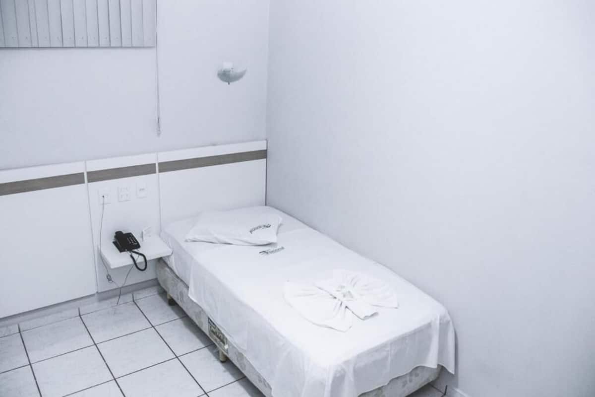 A small, single-bed room with a white bedspread, a white pillow, and a neatly folded white towel. There is a telephone on a small table beside the bed, and a wall-mounted light fixture above the bed.