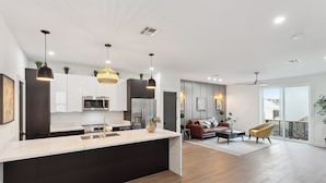 A modern kitchen with a central island, built-in appliances, pendant lighting, and a view to a living area with a sofa and chairs.