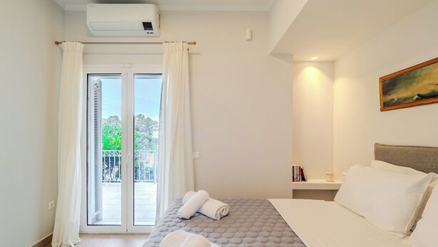 Ein Schlafzimmer mit einem Bett, einem Nachttisch mit Büchern, einem Gemälde an der Wand und einem Balkon mit Blick auf Grünflächen.