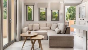 A modern living room with a beige sectional sofa, a round wooden coffee table, and large windows offering a view of greenery outside.