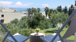 A balcony with two white chairs and a small table, overlooking a coastal area with buildings and greenery.