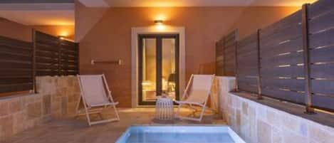 An indoor swimming pool with two lounge chairs, a stone wall, and a door leading to another room.