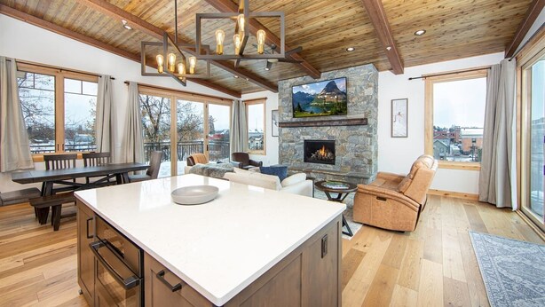A spacious living area with a stone fireplace, wooden ceiling, and large windows offering views of the outdoors. - Luxurious Downtown Penthouse w/ Hot Tub! (Whitefish)