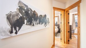 A hallway with a large abstract painting, wooden flooring, and a view into a bedroom with a bed and a window. - Luxurious Downtown Penthouse w/ Hot Tub! (Whitefish)