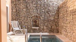 Zona de piscina interior con paredes de piedra, una piscina rectangular, una pequeña ventana de arco y una silla de descanso.