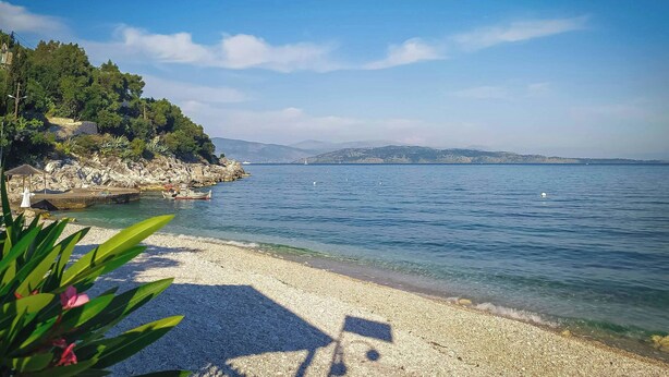 A pebble beach with a clear view of the sea, a rocky outcrop with vegetation, and a distant mountain range. - Villa Delphine by Tsiolis family ★ shared pool (Katavolos)