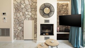 A living room with a stone wall, a fireplace, a flat-screen TV, and two small round tables. - Achileio Alexandros Cottage (Gastouri)