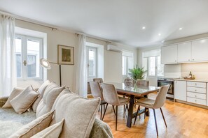Un salon moderne comprenant un canapé, une table à manger et des chaises, un coin cuisine avec des placards et des appareils électroménagers, et de grandes fenêtres laissant entrer la lumière naturelle.