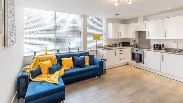 A modern living room with a sofa, a dining area with a kitchen, and large windows with blinds. - Bright 1-Bedroom Apartment with Free Parking (London)