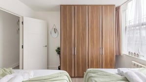 A bedroom with two beds, a wardrobe, a window with sheer curtains, and a wall-mounted clock. - Bright 1-Bedroom Apartment with Free Parking (London)